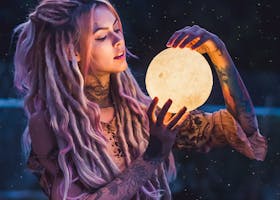 A whimsical outdoor portrait of a tattooed woman holding a glowing orb, conveying mystique and fantasy.